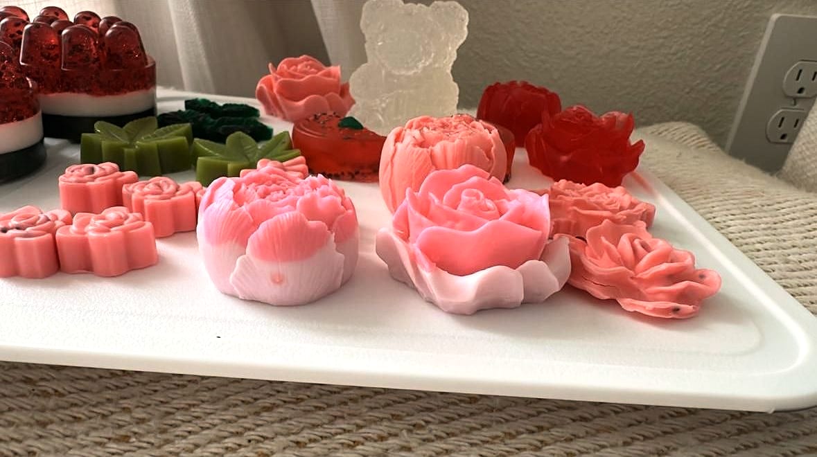 Jabones en forma de rosa, peonía y hojas sobre una bandeja blanca, con un oso transparente y jabones rojos al fondo.