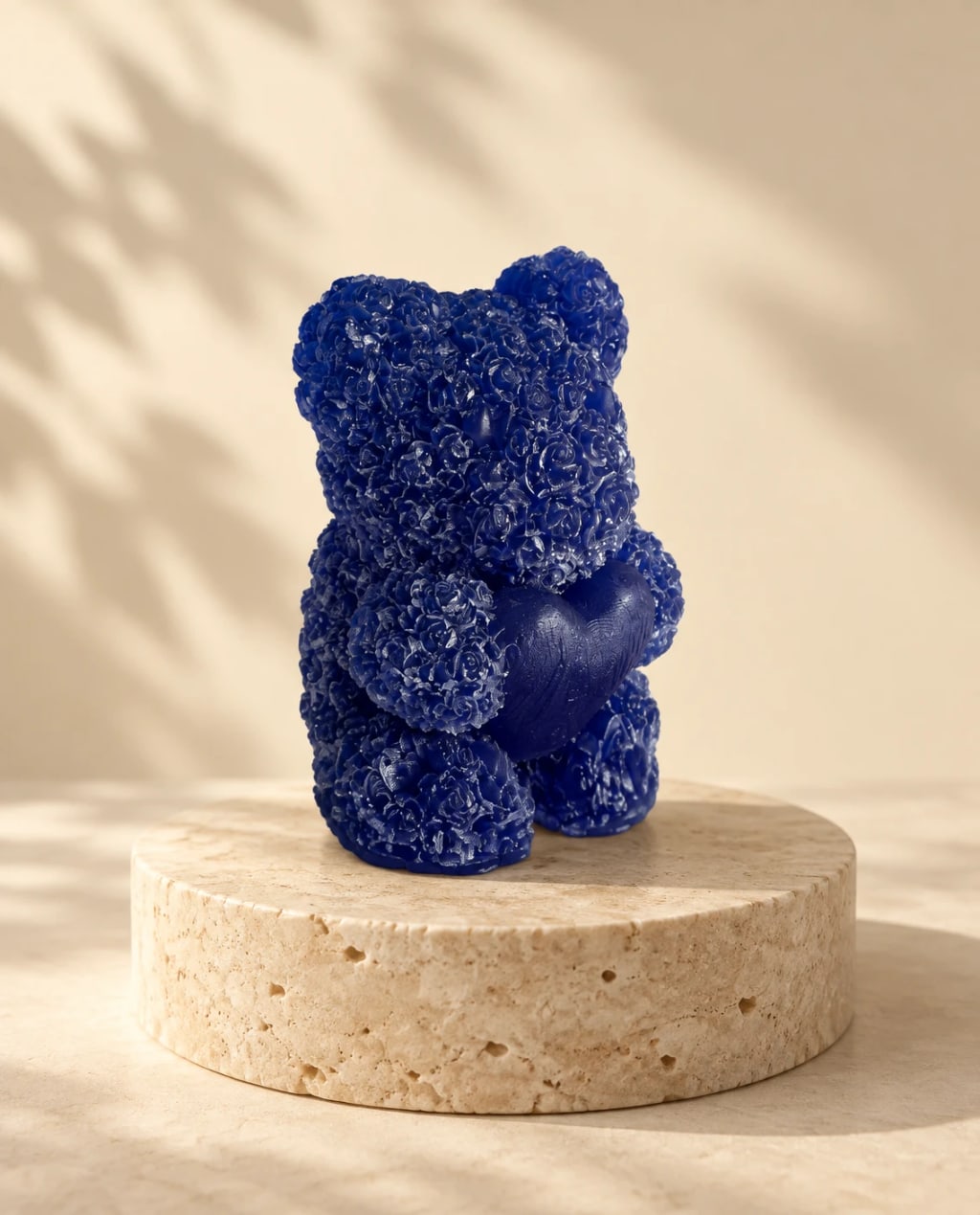 Oso de jabón azul índigo con textura de rosas, sobre un podio de travertino con sombra de hojas; imagen de estudio hecha a partir de la foto real.