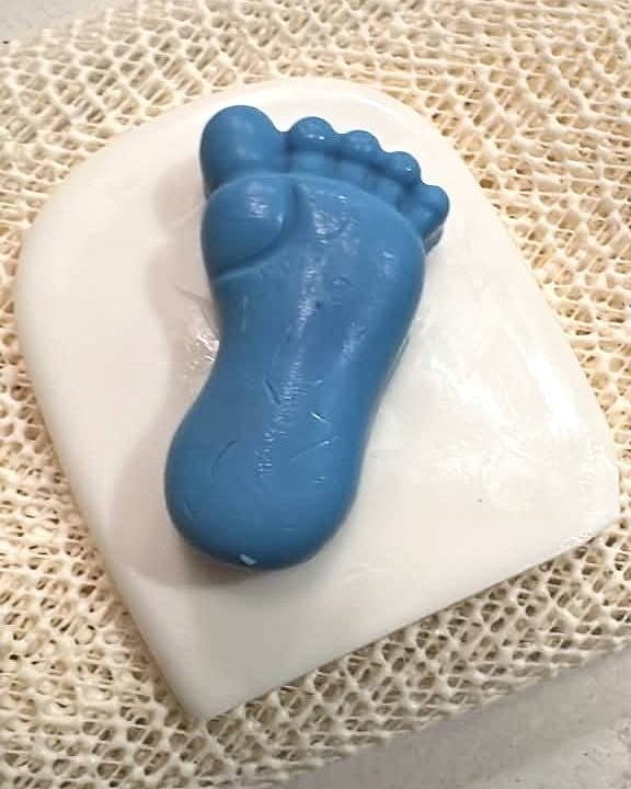 Jabón azul en forma de pie de bebé sobre una base blanca y una malla de lino beige.