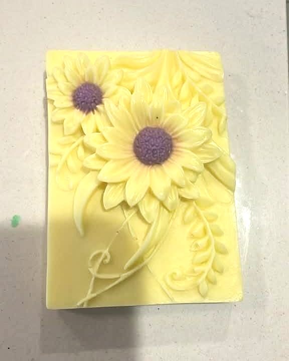 Jabón rectangular en relieve color amarillo con dos girasoles de centro morado y hojas, sobre una superficie gris.